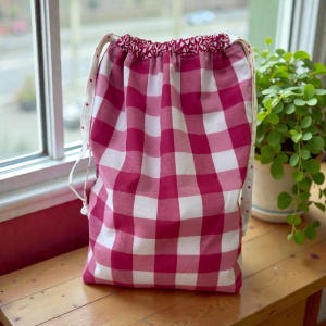 Pode incluir: Uma bolsa com cord&atilde;o com um padr&atilde;o xadrez rosa e branco. A bolsa tem cord&otilde;es brancos e um topo estampado. A bolsa est&aacute; em uma superf&iacute;cie de madeira perto de uma janela e uma planta em vaso.