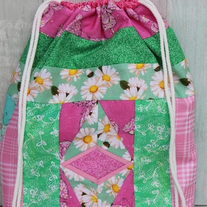 May include: A pink and green drawstring bag with a patchwork design featuring daisies, butterflies, and glitter. The bag has white drawstrings.
