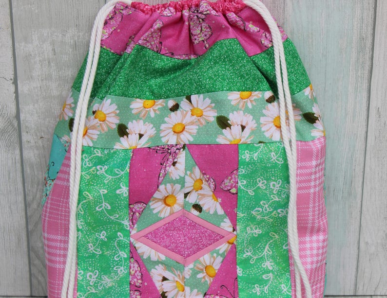 May include: A pink and green drawstring bag with a patchwork design featuring daisies, butterflies, and glitter. The bag has white drawstrings.