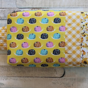 May include: A yellow fabric pouch with a zipper closure and a handle. The pouch is made of patchwork fabric with a pattern of snails, gingham, and floral designs.