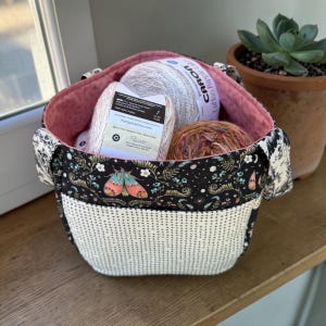 May include: A fabric yarn basket with a black and white floral pattern, a dotted white bottom, and a pink interior. The basket contains several balls of yarn. A moth graphic is on the front of the basket.