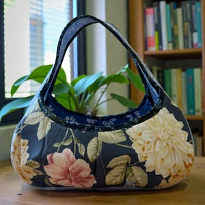 May include: A floral print handbag with a curved handle. The bag features a dark blue background with large cream and pink flower patterns and green leaves. The interior is a solid dark blue. The bag is sitting on a wooden surface.