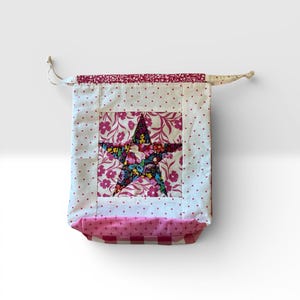Pode incluir: Uma bolsa de tecido artesanal com um desenho de estrela. A estrela &eacute; feita de tecido floral e est&aacute; centrada em um quadrado branco. A bolsa tem um padr&atilde;o de bolinhas rosa e uma parte inferior xadrez rosa e branca.