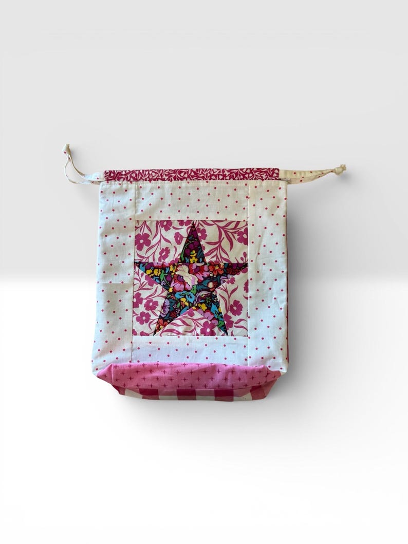 Pode incluir: Uma bolsa de tecido artesanal com um desenho de estrela. A estrela &eacute; feita de tecido floral e est&aacute; centrada em um quadrado branco. A bolsa tem um padr&atilde;o de bolinhas rosa e uma parte inferior xadrez rosa e branca.