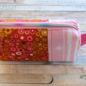 May include: A rectangular zippered pouch with a handle. The pouch is made of fabric with a floral pattern in shades of orange, pink, and brown. The other side has a pink and white striped pattern with a star design. The zipper is silver.