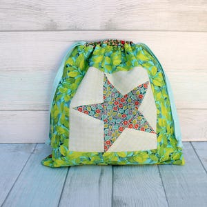 May include: A drawstring bag with a star design. The bag features a green floral pattern with a white star applique. The star is made of a floral fabric with red and blue accents. The drawstring is light blue.