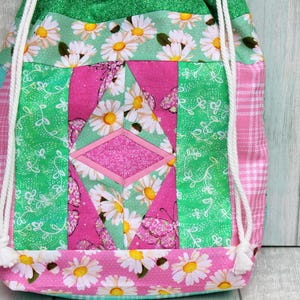 May include: A pink and green drawstring bag with a patchwork design featuring white daisies and butterflies. The bag has white drawstrings.