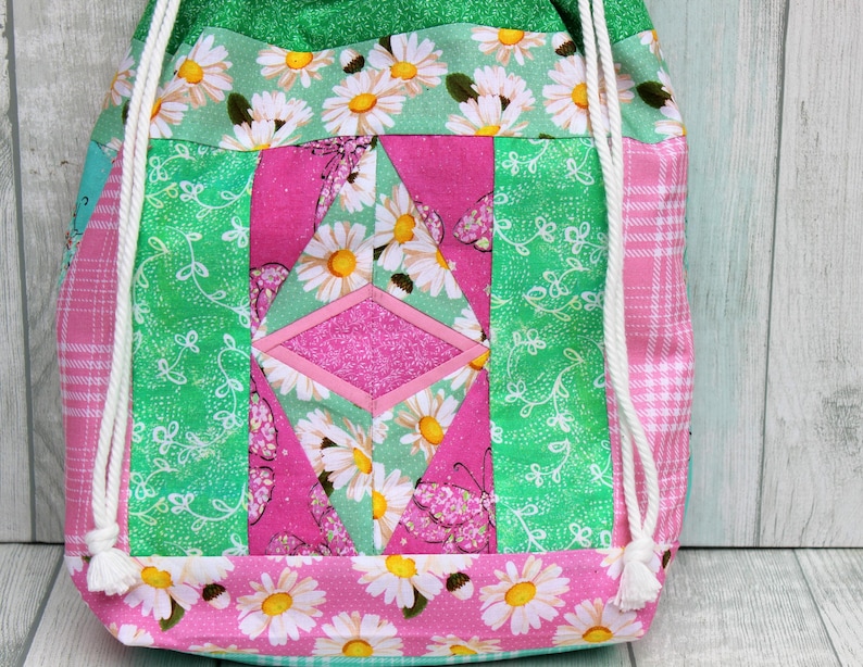 May include: A pink and green drawstring bag with a patchwork design featuring white daisies and butterflies. The bag has white drawstrings.