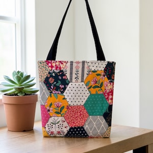 May include: A patchwork tote bag with black handles. The bag's body is composed of various hexagonal fabric pieces in floral and geometric patterns, including yellow, pink, and teal. The bag is sitting on a wooden surface.