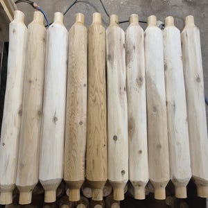 May include: A collection of eleven unfinished wooden posts, varying in shades of light brown and beige. Each post has a cylindrical body with a slightly tapered top and bottom. The wood grain is visible, and some posts have knots.