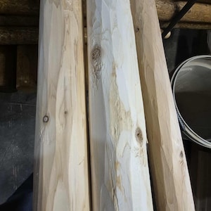 May include: A close-up of several unfinished wooden posts. The posts are light-colored with visible wood grain and knots. The posts have been cut with angled edges. The background is a dark, rustic interior.