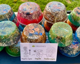 May include: A collection of colorful orgone pyramids with various hues, including blue, green, red, and brown. Each pyramid is filled with metallic flakes and other materials. A small card with diagrams is in the foreground.