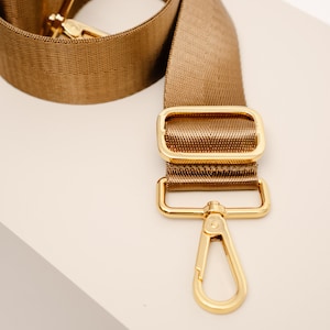 Könnte beinhalten: Ein verstellbarer brauner Gurt mit goldenen Beschlägen, darunter ein Drehverschluss und zwei rechteckige Schlaufen.