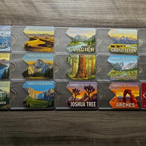 May include: A set of rectangular magnets showcasing various U.S. National Parks. Each magnet depicts a different landscape, such as Crater Lake, Glacier, Grand Teton, and Joshua Tree, with the words "America the Beautiful".