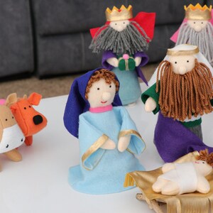 May include: A handmade nativity scene with felt figures. The scene includes figures of the three wise men, Mary, Joseph, baby Jesus, and animals. The figures are in various colors, including blue, purple, green, and gold.