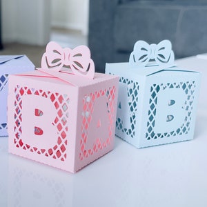 May include: Three small, decorative boxes with cut-out designs. The boxes are in shades of blue, pink, and purple. Each box has a letter cut out of the front, spelling out "BABY".