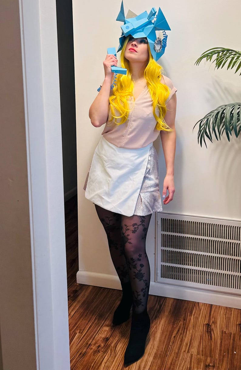 May include: A woman wearing a white skirt, black tights with a floral pattern, and black heeled boots. She is holding a blue phone to her ear and wearing a blue paper hat shaped like a phone.