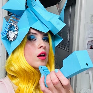 May include: A person with bright yellow hair is wearing a blue paper mache hat shaped like a rotary phone. The hat has a dial with numbers 1-9 and 0. The person is holding a blue paper mache phone to their ear.