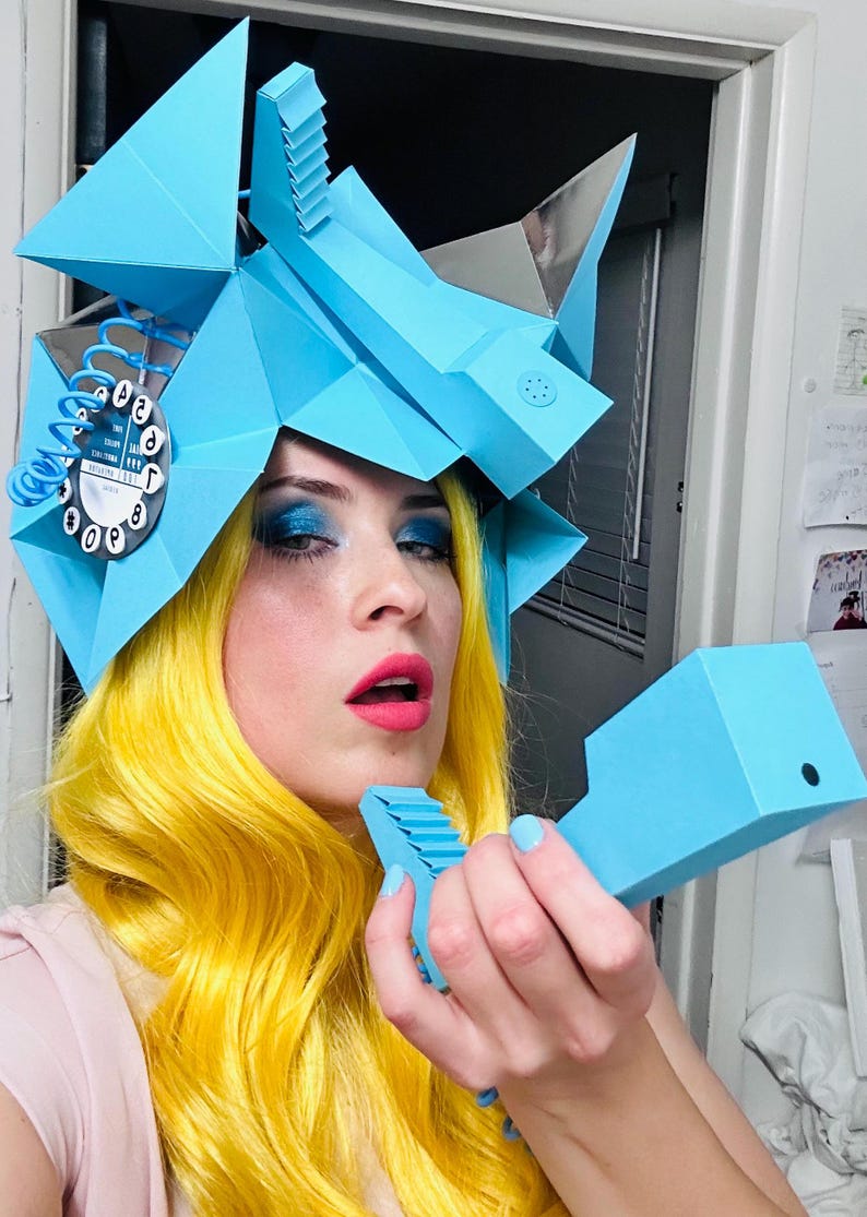 May include: A person with bright yellow hair is wearing a blue paper mache hat shaped like a rotary phone. The hat has a dial with numbers 1-9 and 0. The person is holding a blue paper mache phone to their ear.