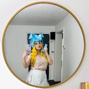 May include: A person wearing a blue paper hat and a pink shirt is holding a white phone and taking a selfie in a mirror.