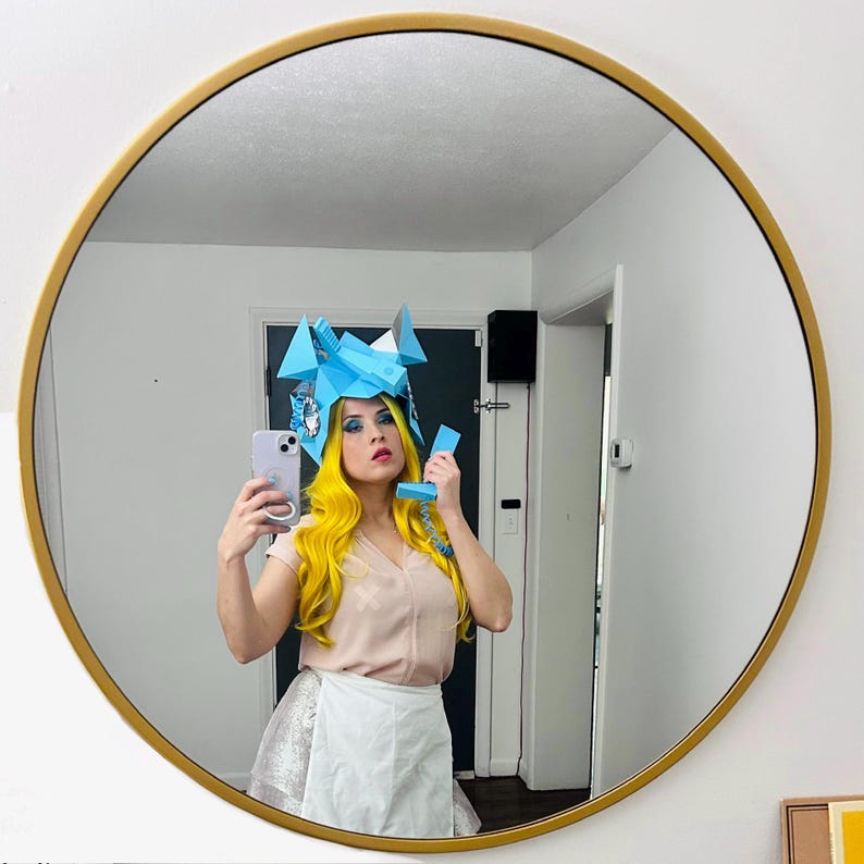 May include: A person wearing a blue paper hat and a pink shirt is holding a white phone and taking a selfie in a mirror.