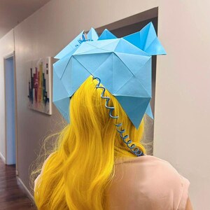 May include: A person with long, bright yellow hair wearing a blue origami hat with a blue coiled wire attached to the back.