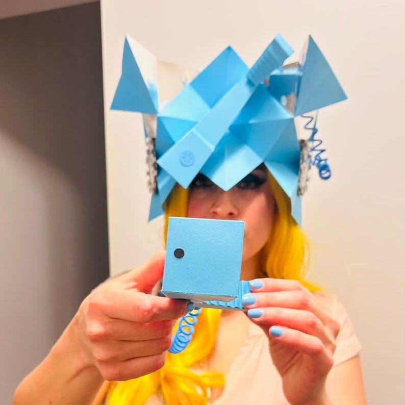 May include: A person wearing a blue paper crown with a blue paper box in their hands. The box has a black dot on the front.
