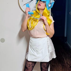 May include: A woman with bright yellow hair is wearing a blue and white paper hat shaped like a rotary phone. She is holding a blue rotary phone receiver to her ear and smiling.