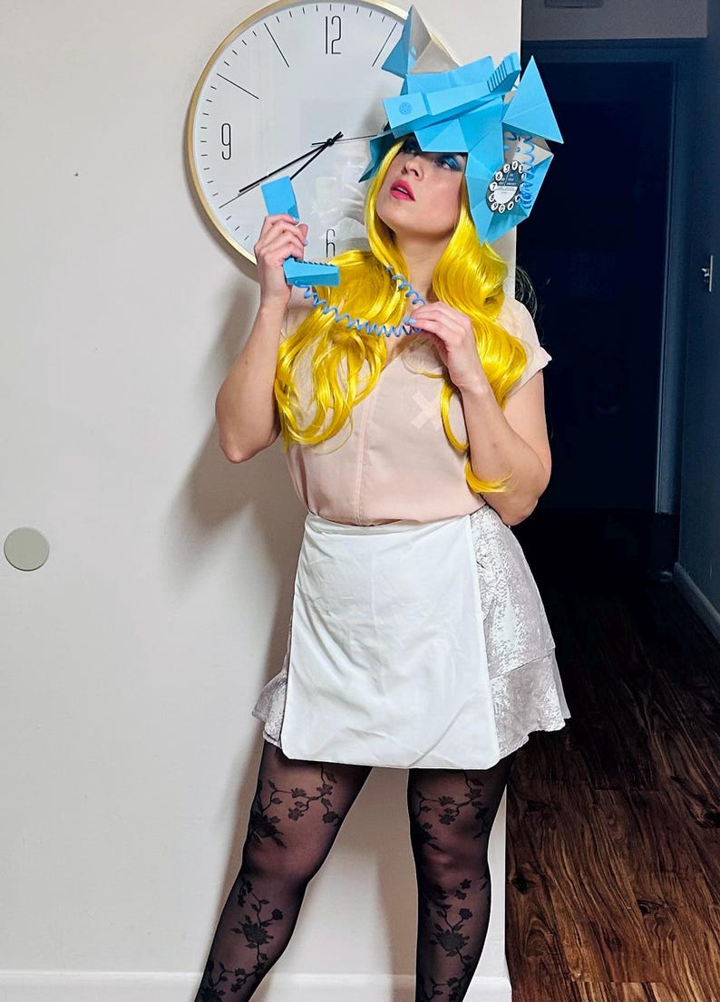 May include: A woman with bright yellow hair is wearing a blue and white paper hat shaped like a rotary phone. She is holding a blue rotary phone receiver to her ear and smiling.