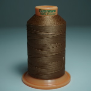 Puede incluir: Una bobina de hilo marrón Gutermann, etiquetada "100% Poliéster", en una bobina de plástico.