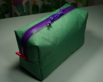 May include: A green zippered pouch with a purple zipper and a pink pull tab. The pouch is made of a textured fabric.