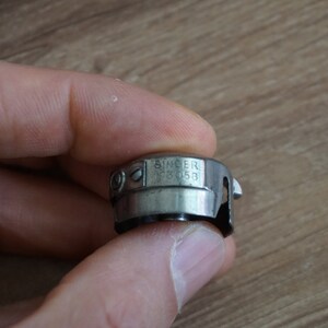 Pu&ograve; includere: Un portabobina per macchina da cucire Singer, modello FB 053, in metallo color argento. Il portabobina &egrave; tenuto in mano su uno sfondo sfocato in legno. Il logo Singer &egrave; visibile.