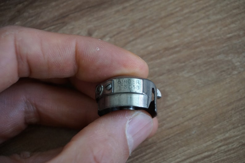 Pu&ograve; includere: Un portabobina per macchina da cucire Singer, modello FB 053, in metallo color argento. Il portabobina &egrave; tenuto in mano su uno sfondo sfocato in legno. Il logo Singer &egrave; visibile.