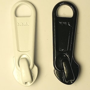 以下が含まれることがあります： 白と黒の2つのファスナー引き手のクローズアップ。白い引き手には「YKK」の文字が刻印されています。どちらの引き手も、持ちやすいように丸みを帯びた上部と、ファスナーに取り付けるための湾曲した底部を備えています。