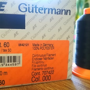 Puede incluir: Una bobina de hilo negro con una etiqueta naranja. La etiqueta dice "No./Tkt. 60 dtex 500(3)/tex 50 0542121 Made in Germany 100% POLYESTER Continuous Filament Endloser Nähfaden Fil continu Filo continuo Filamento continuo Cont. 707422 Col. 000 Tera 60 10 x 600 m".