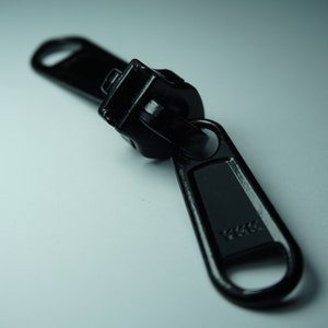 Pode incluir: Um puxador de zíper de metal preto com o logotipo YKK. O puxador de zíper é projetado para ser usado com um zíper em uma roupa ou bolsa.