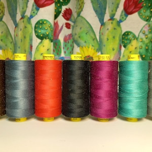 以下が含まれることがあります： 茶色、ティール、赤、黒、マゼンタ、ターコイズ、グレーの色合いのカラフルな糸巻きの列。 スプールは円筒形で、黄色のラベルが付いています。 背景にはサボテンをテーマにした生地があります。