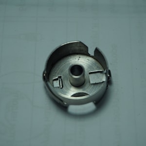 Pu&ograve; includere: Un portabobina per macchina da cucire color argento. La cassa &egrave; circolare con un foro centrale e varie fessure e linguette sul bordo. Il metallo ha una superficie leggermente riflettente e il design &egrave; funzionale e industriale.