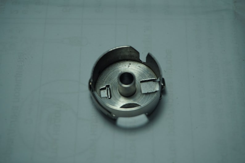 Pu&ograve; includere: Un portabobina per macchina da cucire color argento. La cassa &egrave; circolare con un foro centrale e varie fessure e linguette sul bordo. Il metallo ha una superficie leggermente riflettente e il design &egrave; funzionale e industriale.
