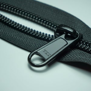 May include: A close-up of a black zipper with a black metal slider. The slider has the word "YKK" printed on it.