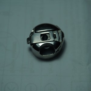 Pu&ograve; includere: Un portabobina lucido e metallico per macchina da cucire. L'involucro ha una forma arrotondata con un fermo a cerniera e un'apertura centrale. &Egrave; progettato per contenere una bobina di filo per macchine da cucire.