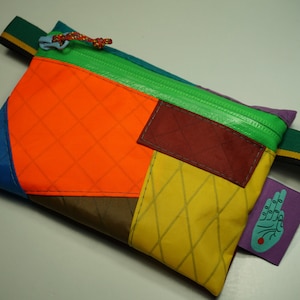 May include: A small, multi-colored zippered pouch with a diamond pattern. The pouch is made of different fabrics in shades of blue, green, orange, yellow, brown, and red. A small patch with a hand design is attached to the side.