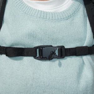 Può includere: Primo piano di una cinghia pettorale per zaino nera con fibbia in plastica. La cinghia è attaccata a uno zaino nero con il logo Lowe Alpine. La persona indossa un maglione azzurro.