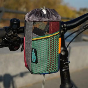 Op de afbeelding: Een veelkleurige fietstas voor het stuur met een trekkoordsluiting. De tas heeft een patchwork-ontwerp met grijze, groene, rode en geruite stoffen. Een groen mesh-vak en een oranje accent zijn zichtbaar aan de voorkant. Het is bevestigd aan een zwart stuur.