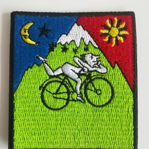 May include: An embroidered patch featuring a psychedelic design with a person riding a bicycle on a green hill. The background is divided into a blue section with a crescent moon and a red section with a sun. The hill is topped with white mountains.