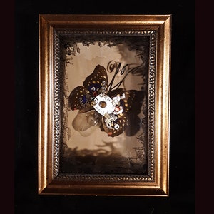 May include: A framed artwork featuring a butterfly made of metal gears and other mechanical parts. The butterfly is set against a dark background and has a light bulb in the center.