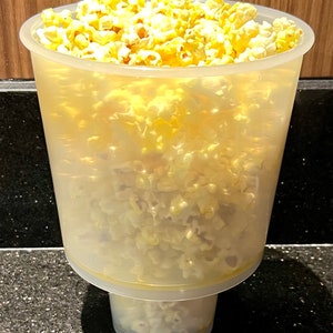 May include: A clear plastic popcorn container filled with freshly popped, yellow popcorn. The container has a wide top and a smaller, attached base. The popcorn is overflowing, suggesting a generous serving.