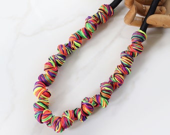 Puede incluir: Un collar colorido hecho de hilo trenzado en tonos de morado, rosa, verde, amarillo y negro. El collar está encordado en un cordón negro y tiene un diseño anudado.