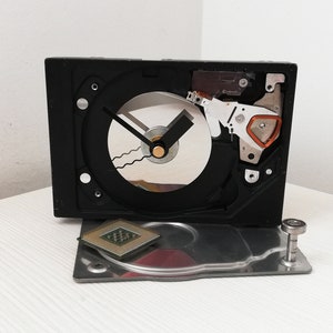 May include: A unique, handmade clock crafted from a repurposed computer hard drive. The black clock features a circular face with black hands and a silver disc. A CPU and metal plate are placed in front of the clock.