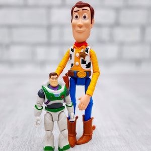 Puede incluir: Figuras de juguete de Woody y Buzz Lightyear de la franquicia Toy Story. Woody lleva su sombrero de vaquero, chaleco y pantalones vaqueros caracter&iacute;sticos. Buzz Lightyear lleva su traje de ranger espacial. Las figuras est&aacute;n de pie sobre una superficie de madera clara.